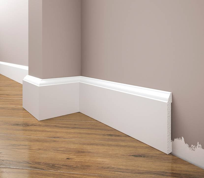 Białe listwy przypodłogowe z frezowanym wzorem, zamontowane w rogu pomieszczenia z drewnianą podłogą, widoczne ślady farby na ścianie.
