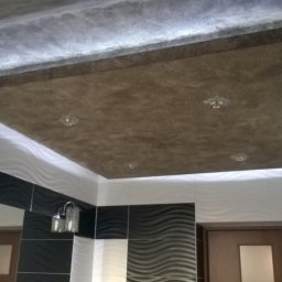 Damian Kwieciński - Sufit w łazience z podwieszanym elementem w kolorze brązowym, oświetleniem punktowym i dekoracyjną fakturą, ściany wykończone płytkami w biało-czarne fale.