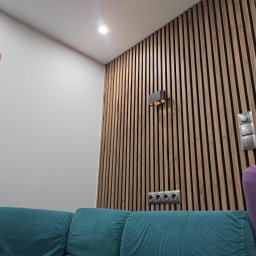 Damian Kwieciński - Drewniane lamele na ścianie z oświetleniem punktowym i gniazdkami, widoczny fragment sofy w odcieniach turkusu i fioletu oraz obraz w złotej ramie.