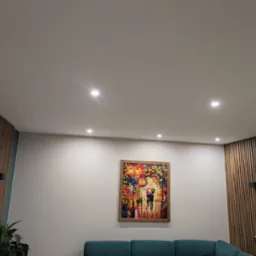 Jasne wnętrze pokoju z gładką, pomalowaną na biało ścianą i sufitem, oświetlonymi punktowymi lampami. Na ścianie wisi kolorowy obraz w drewnianej ramie, a część ściany pokryta jest drewnianymi...