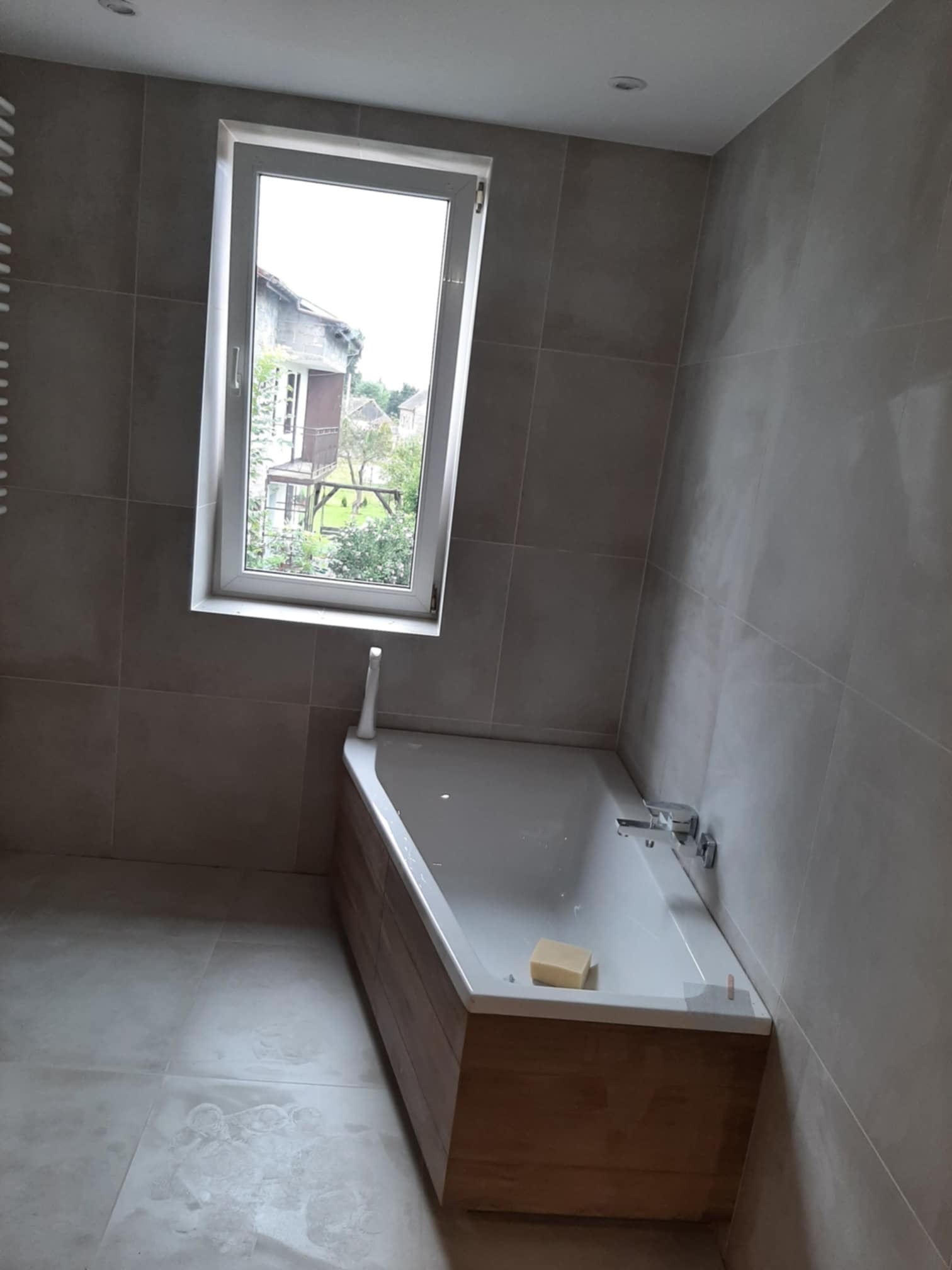 Wykończona łazienka z wanną obudowaną drewnem, szare płytki na ścianach i podłodze, okno z widokiem na zieleń, kostka mydła na brzegu wanny.