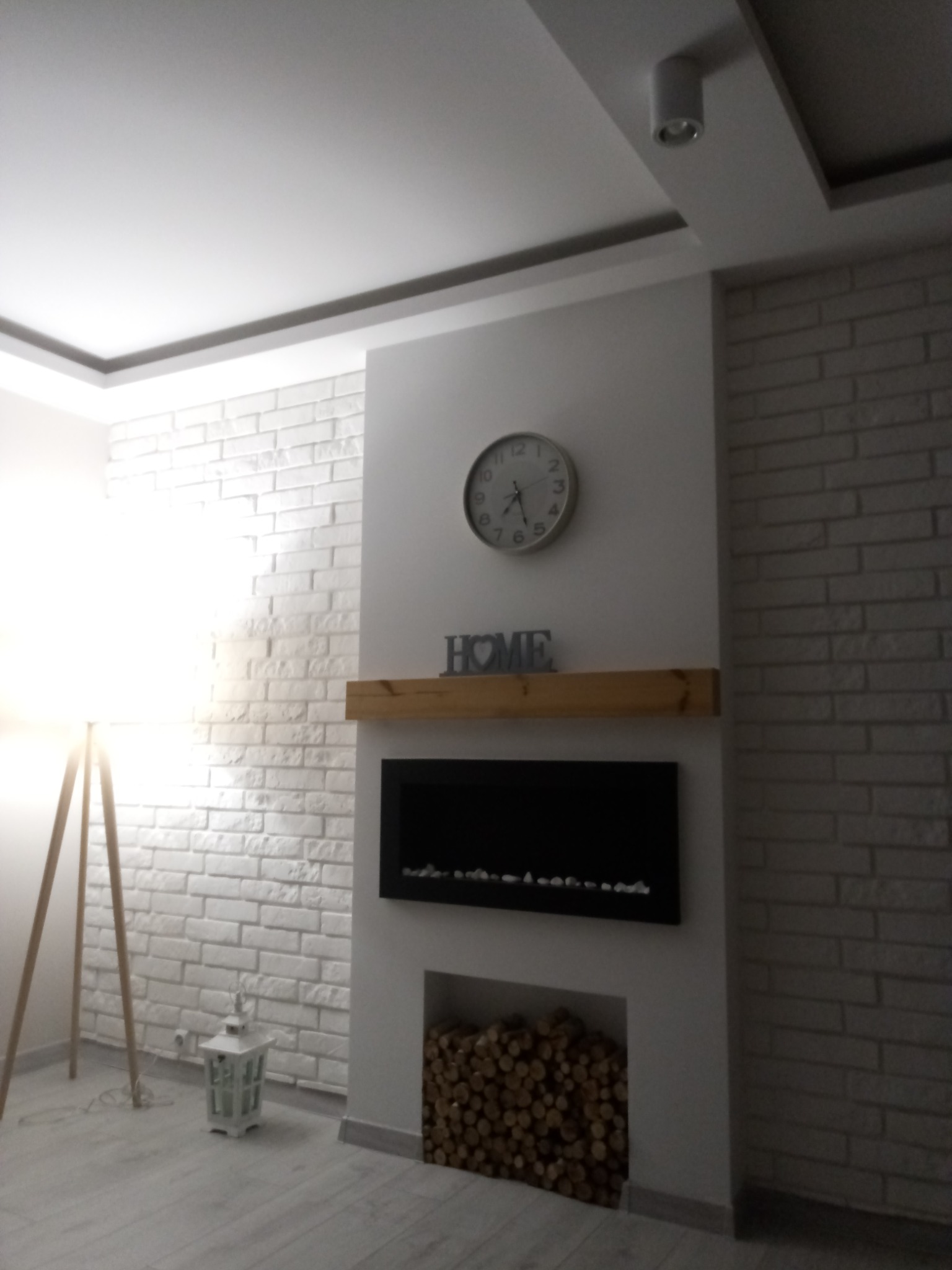 Biała ściana z cegły, kominek elektryczny z drewnianą półką, zegar na ścianie i dekoracje w nowoczesnym salonie z lampą podłogową.