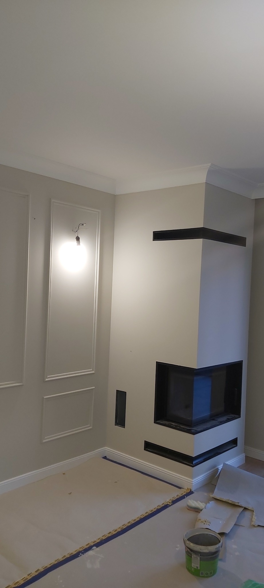 Wnętrze w trakcie prac wykończeniowych: ściany z dekoracyjnymi panelami i nowoczesnym kominkiem z czarnymi wnękami, podłoga zabezpieczona folią i taśmą malarską, widoczne wiadro z farbą.