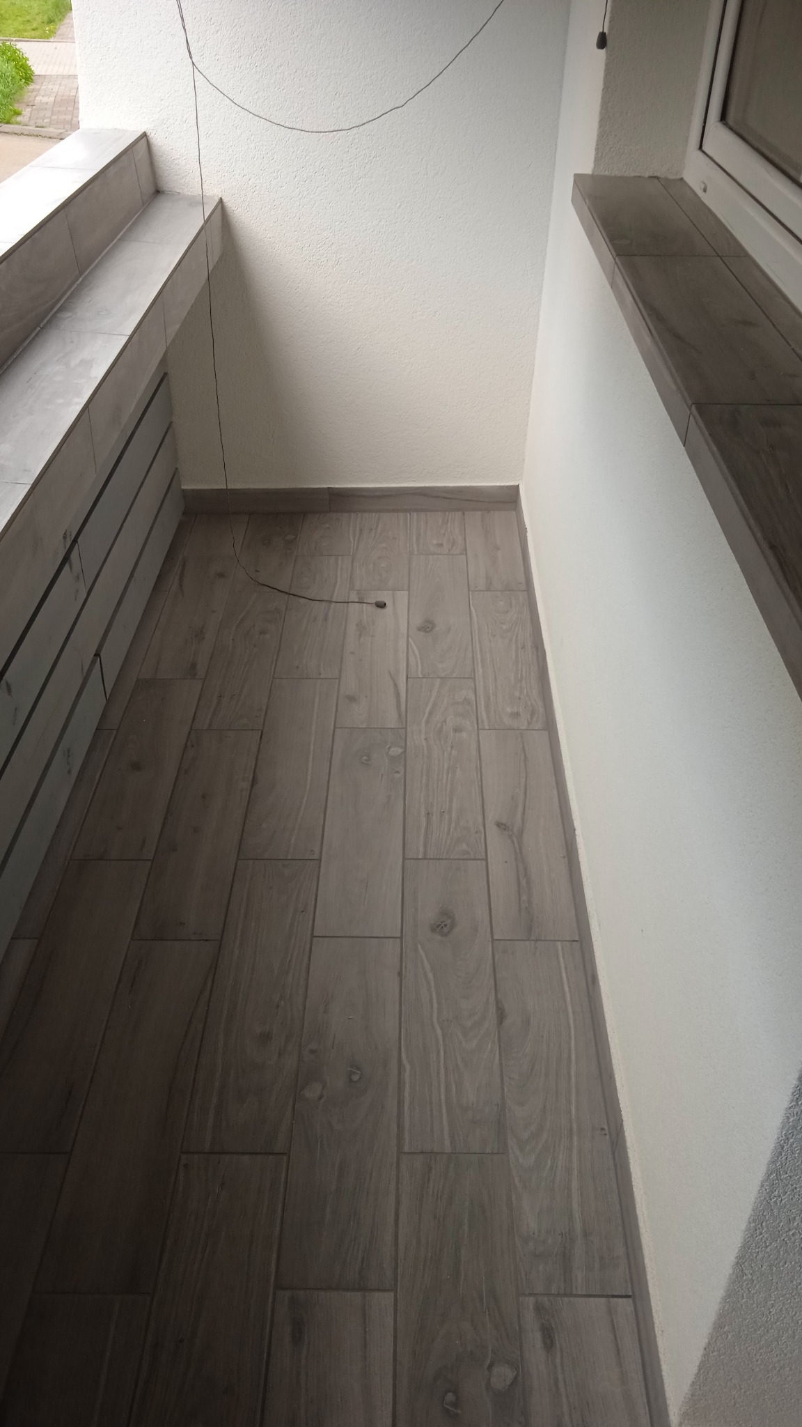 Wykończony balkon z szarymi płytkami imitującymi drewno na podłodze i parapetach, białe ściany o delikatnej strukturze.