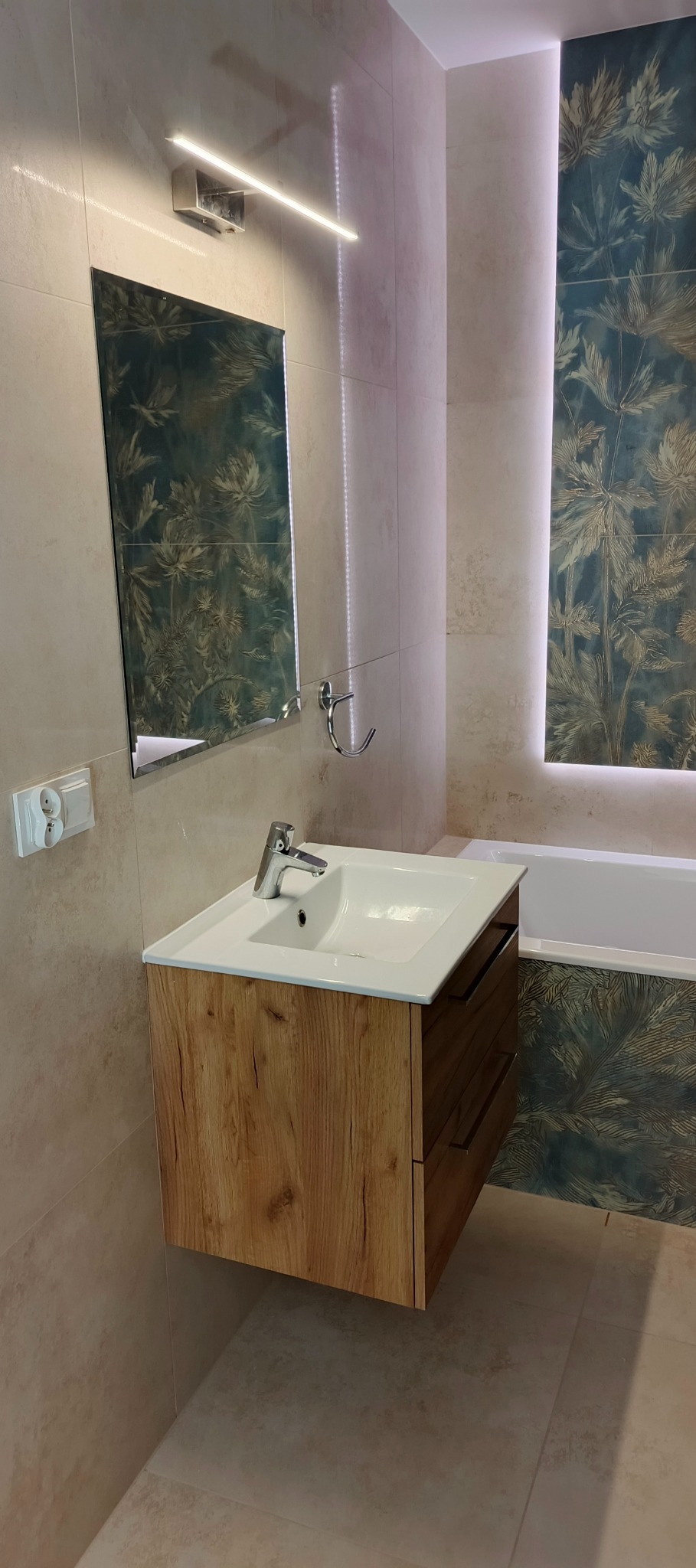 Nowoczesna łazienka z drewnianą szafką pod umywalką, lustrem z podświetleniem LED i dekoracyjną płytką z motywem roślinnym w strefie wanny.