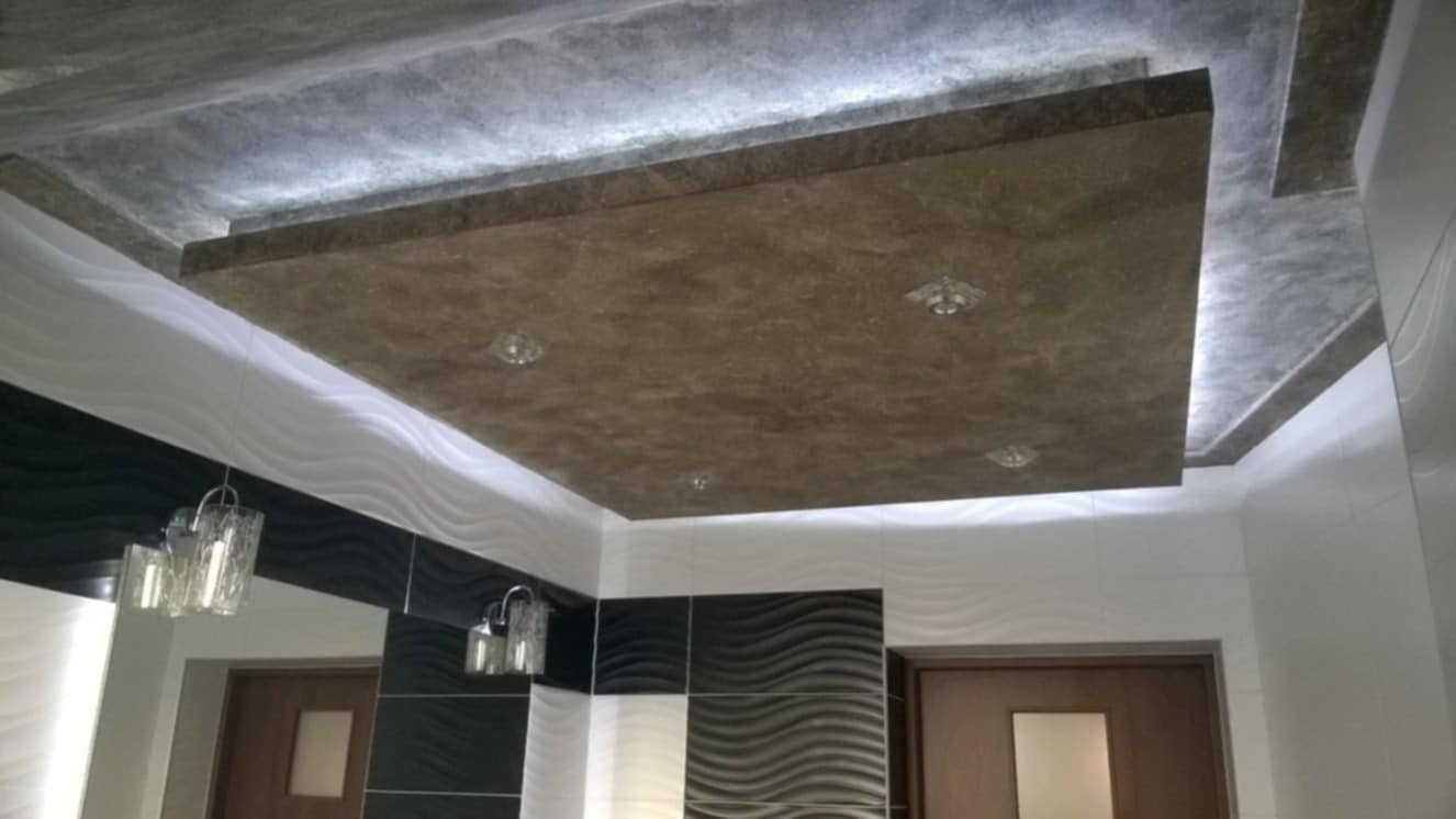 Sufit w łazience z podwieszanym elementem w kolorze brązowym, oświetleniem punktowym i dekoracyjną fakturą, ściany wykończone płytkami w biało-czarne fale.