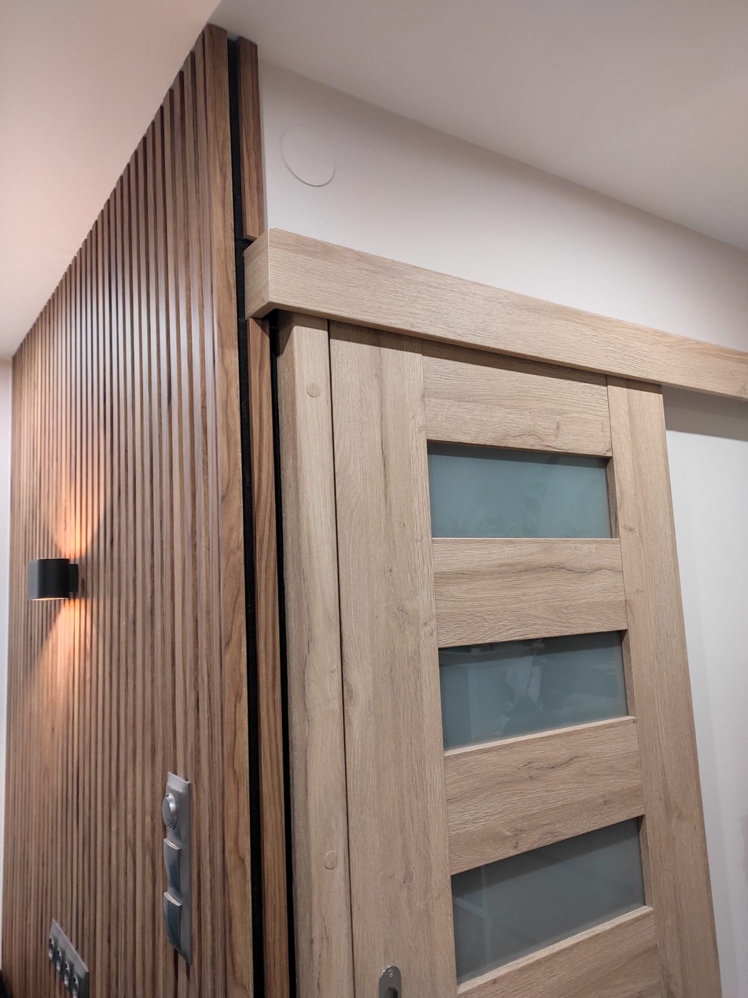 Panel ścienny z drewnianych listewek z czarnym oświetleniem punktowym, sąsiadujący z przesuwnymi drzwiami z jasnego drewna z trzema matowymi szybami.