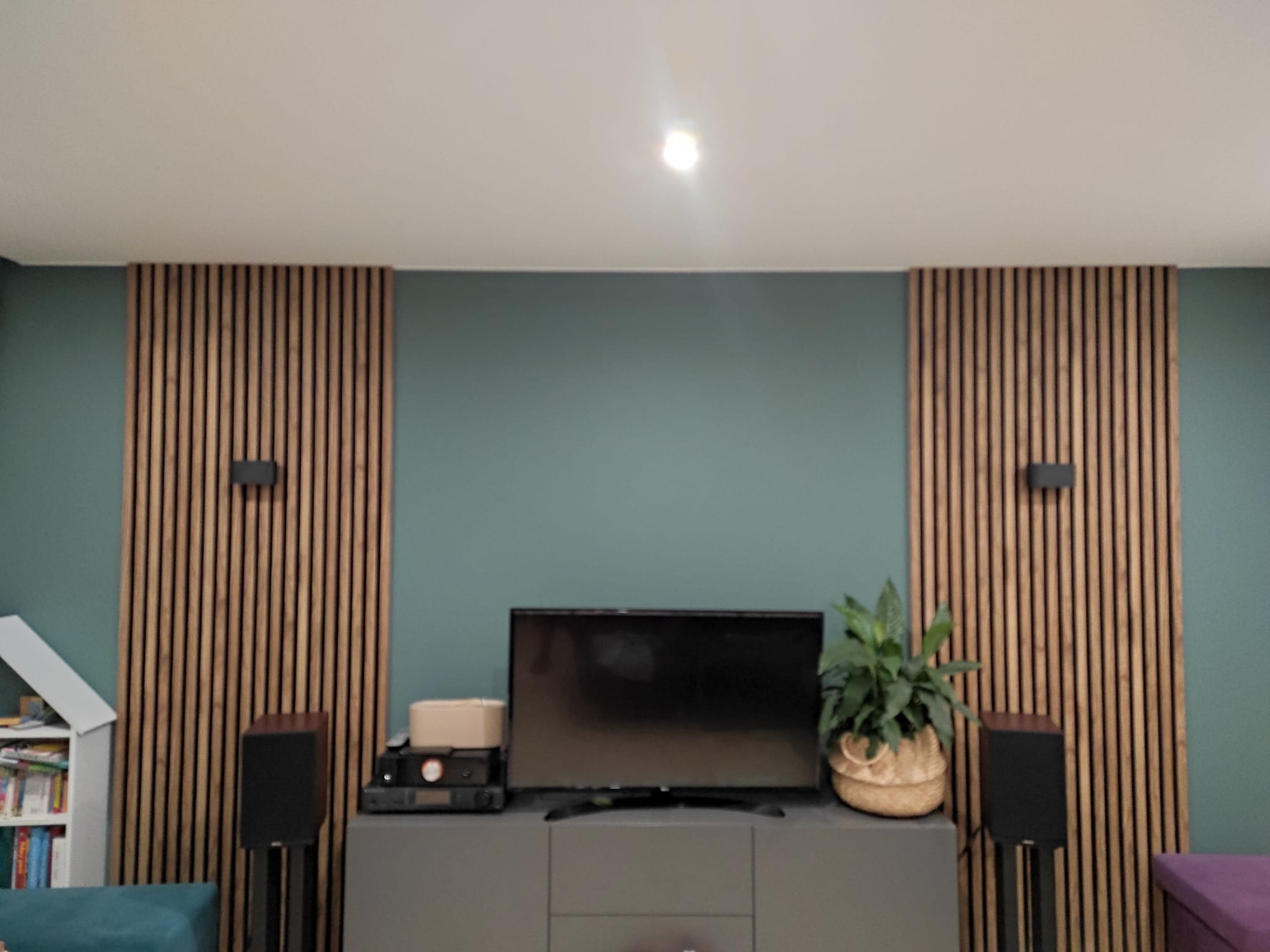 Telewizor i sprzęt audio na szafce RTV na tle ściany z drewnianych paneli dekoracyjnych i zielonej farby.