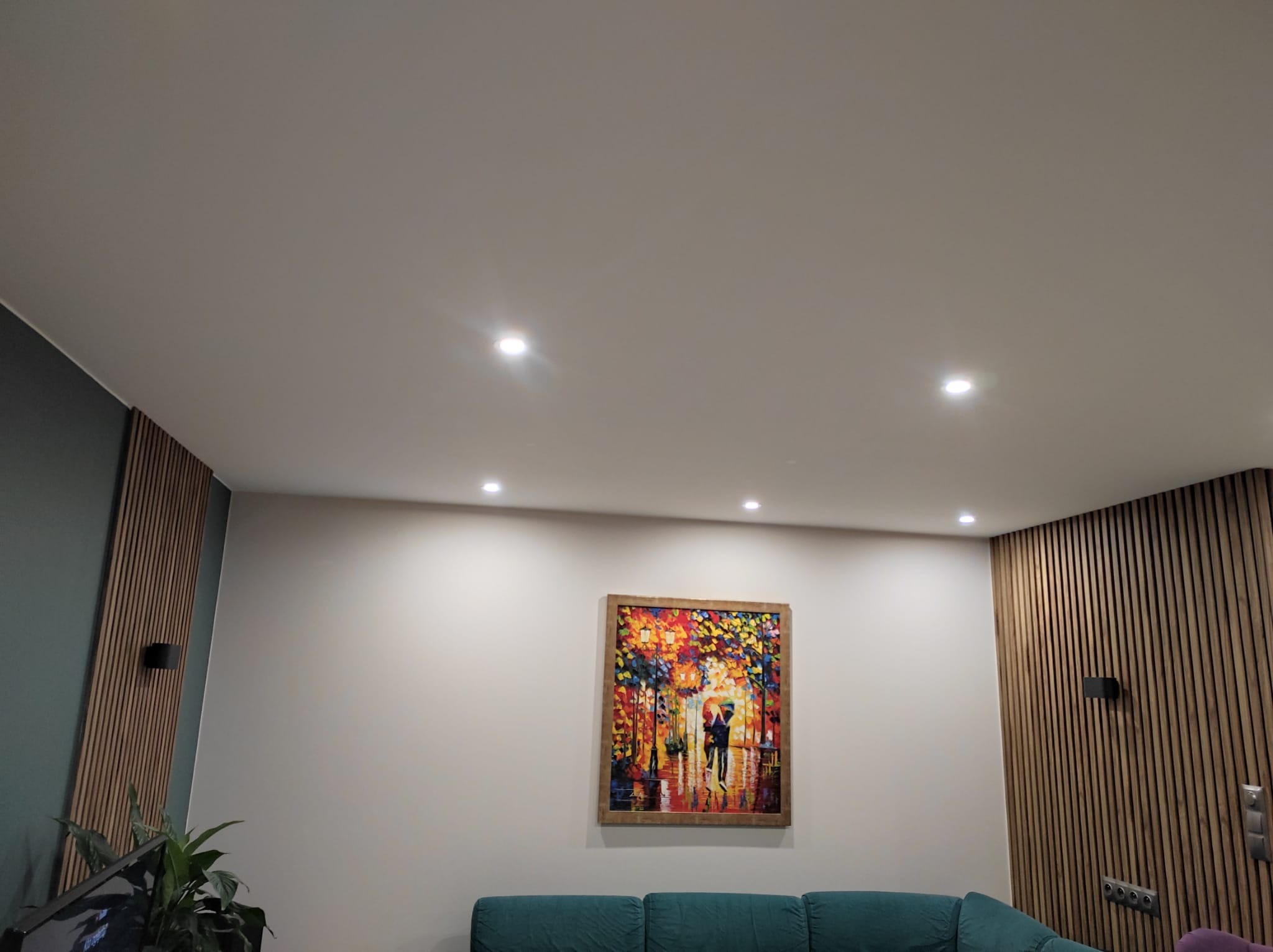 Jasne wnętrze pokoju z gładką, pomalowaną na biało ścianą i sufitem, oświetlonymi punktowymi lampami. Na ścianie wisi kolorowy obraz w drewnianej ramie, a część ściany pokryta jest drewnianymi...