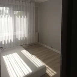 Jasne wnętrze pokoju z drewnianą podłogą, grzejnikiem i białymi zasłonami w oknie, częściowo widoczne łóżko z pościelą w promieniach słońca.