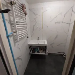 Biała umywalka z szafką w łazience z marmurowymi płytkami, widoczne narzędzia i rury instalacyjne, grzejnik drabinkowy na ścianie.