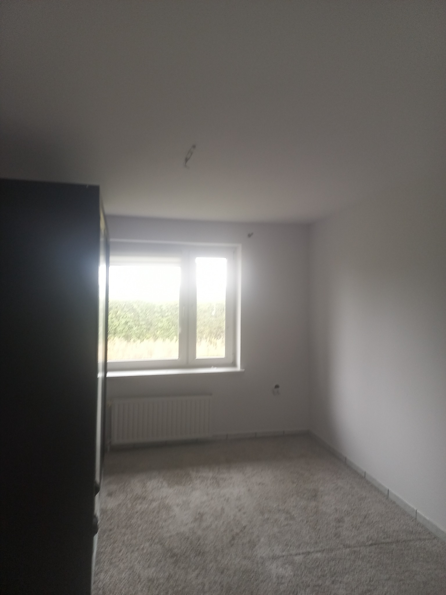 Pusty pokój po remoncie z jasnymi ścianami, nową wykładziną dywanową, grzejnikiem pod oknem i widocznymi śladami po instalacji oświetlenia na suficie.