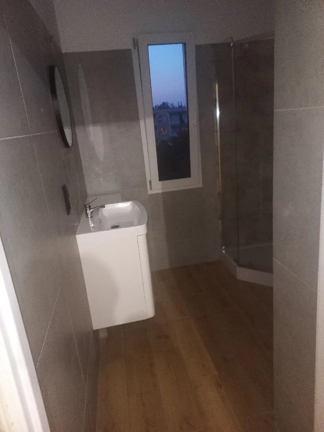 Wykończona łazienka z umywalką na szafce, kabiną prysznicową w rogu i drewnopodobną podłogą, widok przez okno na budynek mieszkalny o zmierzchu.