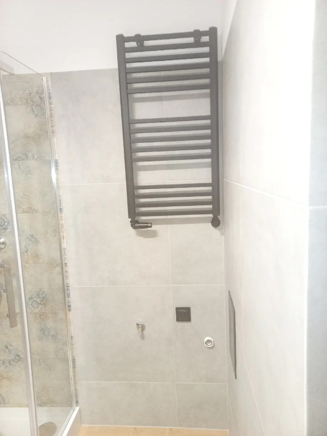 Wykończona łazienka z grzejnikiem drabinkowym w kolorze antracytowym, zamontowanym na jasnoszarych płytkach, obok kabiny prysznicowej z wzorzystymi płytkami wewnątrz.