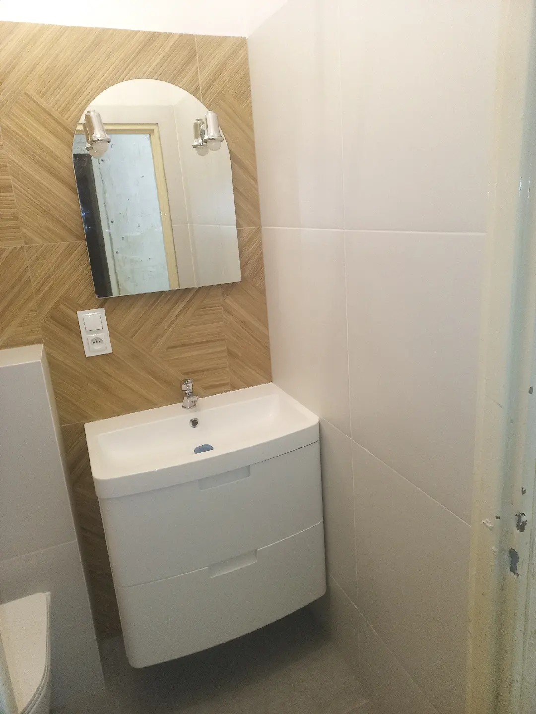 Nowoczesna łazienka po remoncie: biała szafka pod umywalkę z dwoma szufladami, owalne lustro z lampkami na ścianie wyłożonej płytkami imitującymi drewno, białe płytki na sąsiedniej ścianie, szara...