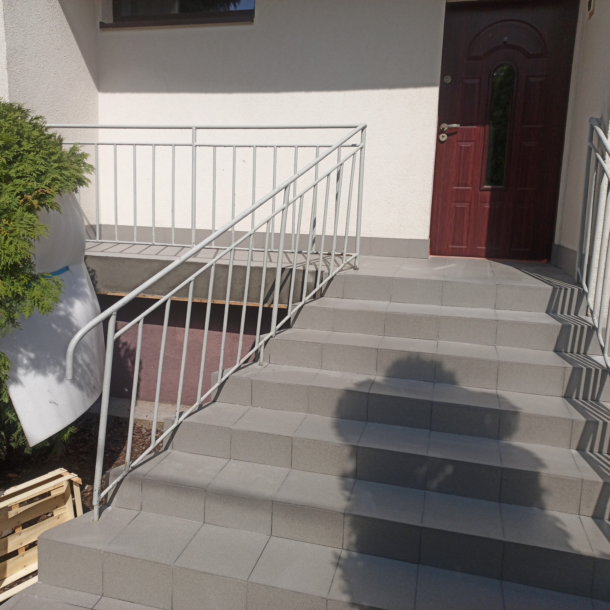 Zewnętrzne schody wejściowe wyłożone szarą, kwadratową terakotą, z metalową, pomalowaną na szaro barierką, prowadzące do ciemnych drzwi wejściowych. Obok schodów widoczny krzew iglasty i drewniana...