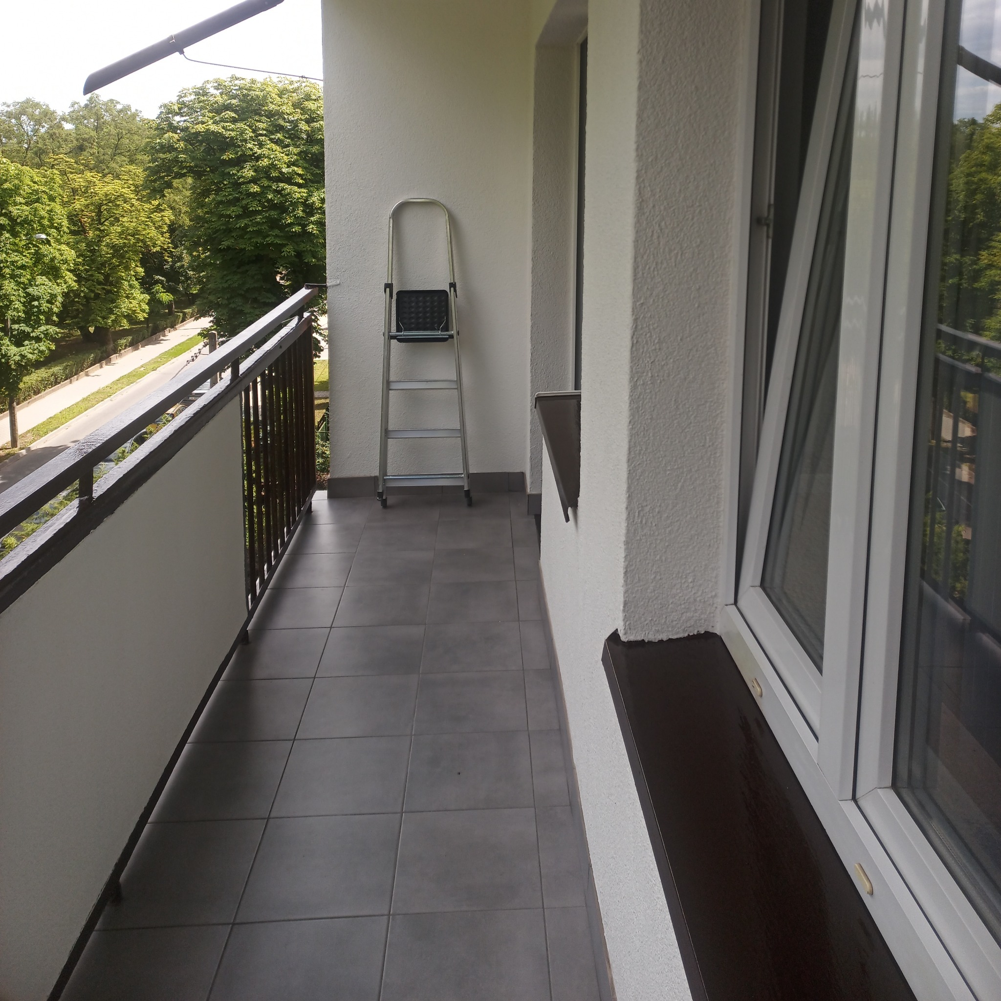 Wykończony balkon z szarymi płytkami podłogowymi, białą balustradą i ścianami o fakturze baranka. Aluminiowa drabina oparta o ścianę. Widok na zieleń drzew za balkonem.