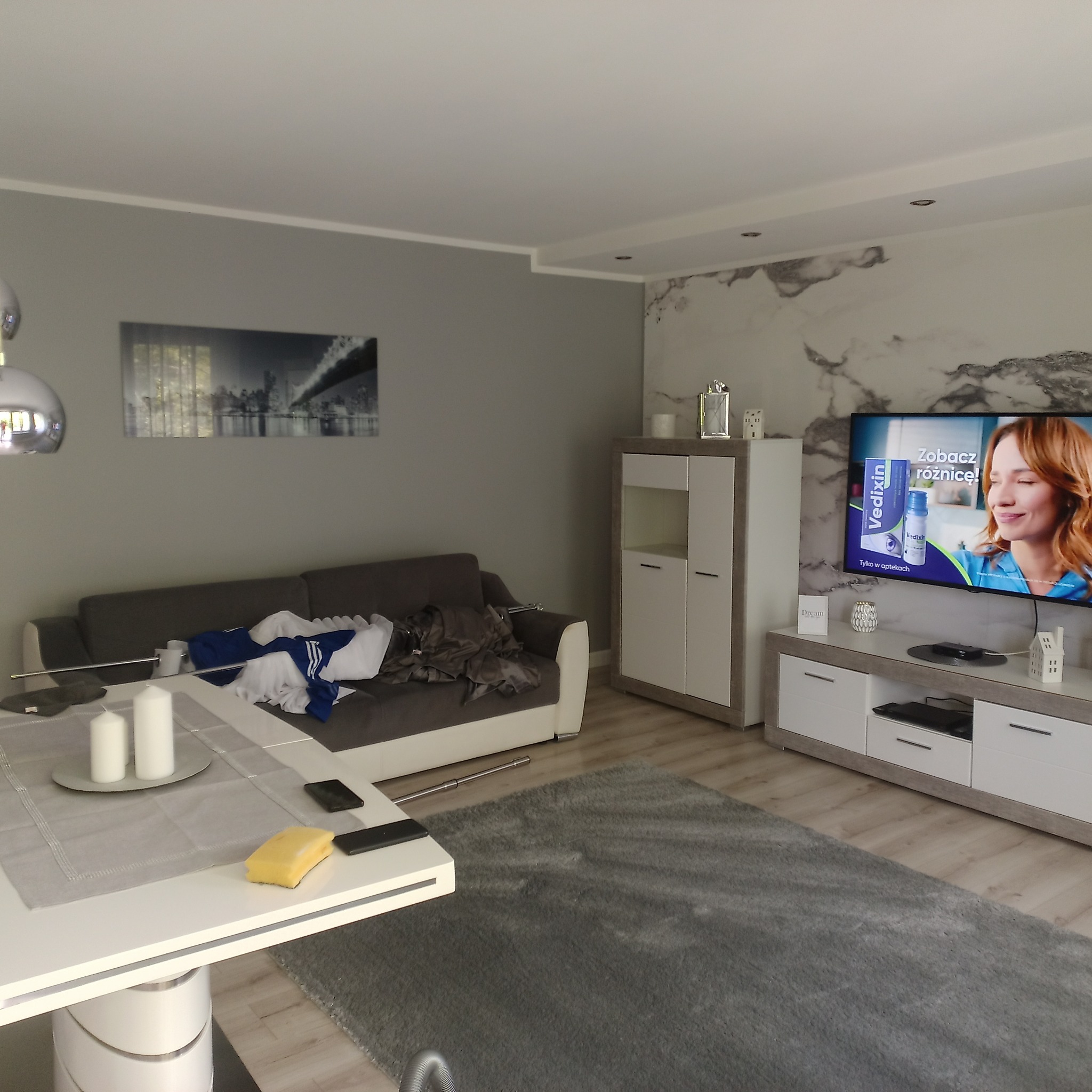 Wnętrze salonu z szarą kolorystyką, widoczna sofa z ubraniami, stolik z dekoracjami i gąbką, telewizor z reklamą leku, szafka RTV i dywan.