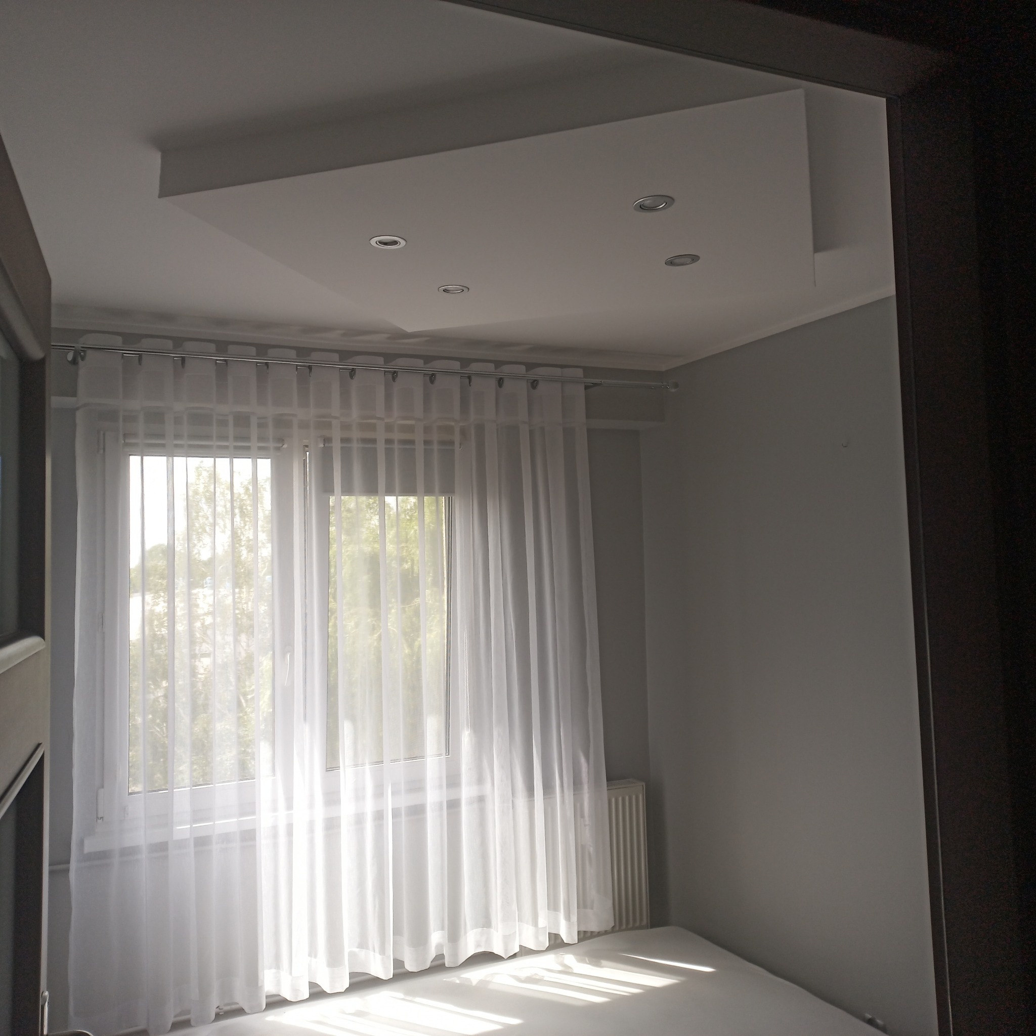 Jasne wnętrze pokoju po odświeżeniu, z szarymi ścianami, białym sufitem z wbudowanymi punktowymi lampami, białymi firanami w oknie i grzejnikiem pod nim.