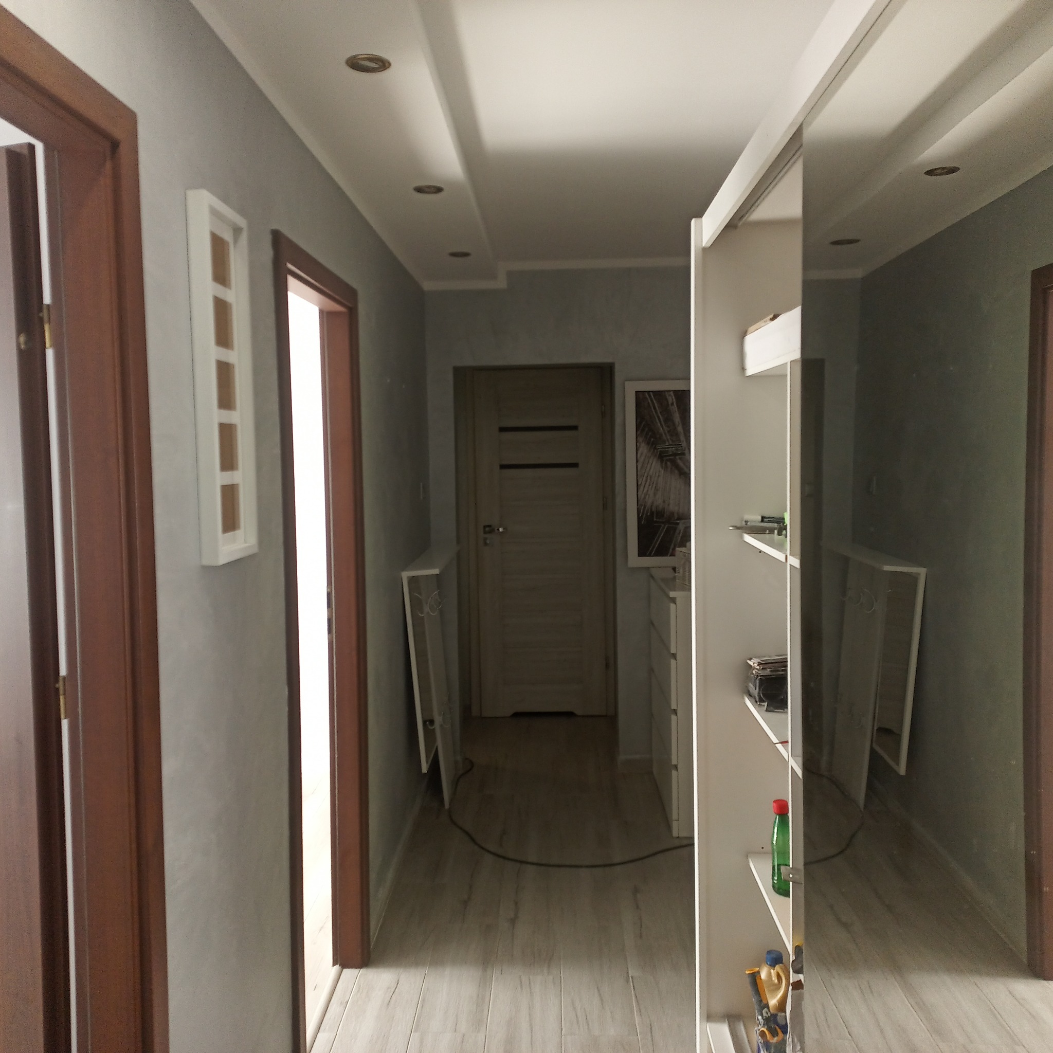 Długi korytarz w mieszkaniu po odświeżeniu, z szarą ścianą, drewnianymi drzwiami, panelami podłogowymi i białą zabudową szafy z lustrzanymi drzwiami.