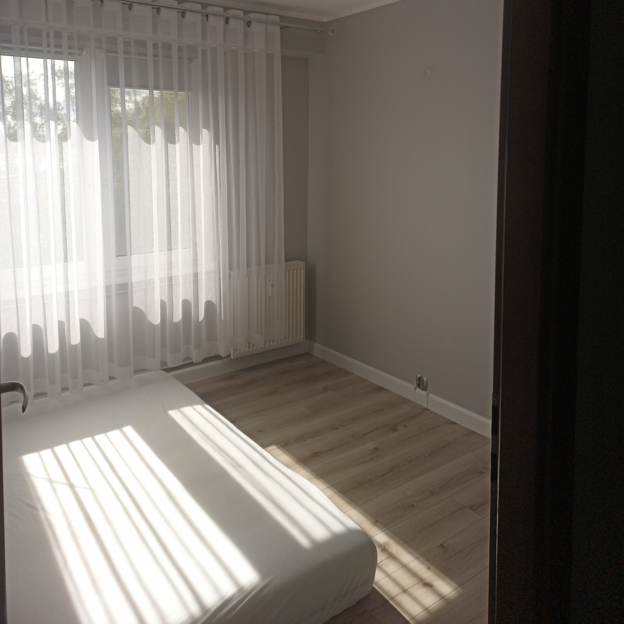 Jasne wnętrze pokoju z drewnianą podłogą, grzejnikiem i białymi zasłonami w oknie, częściowo widoczne łóżko z pościelą w promieniach słońca.