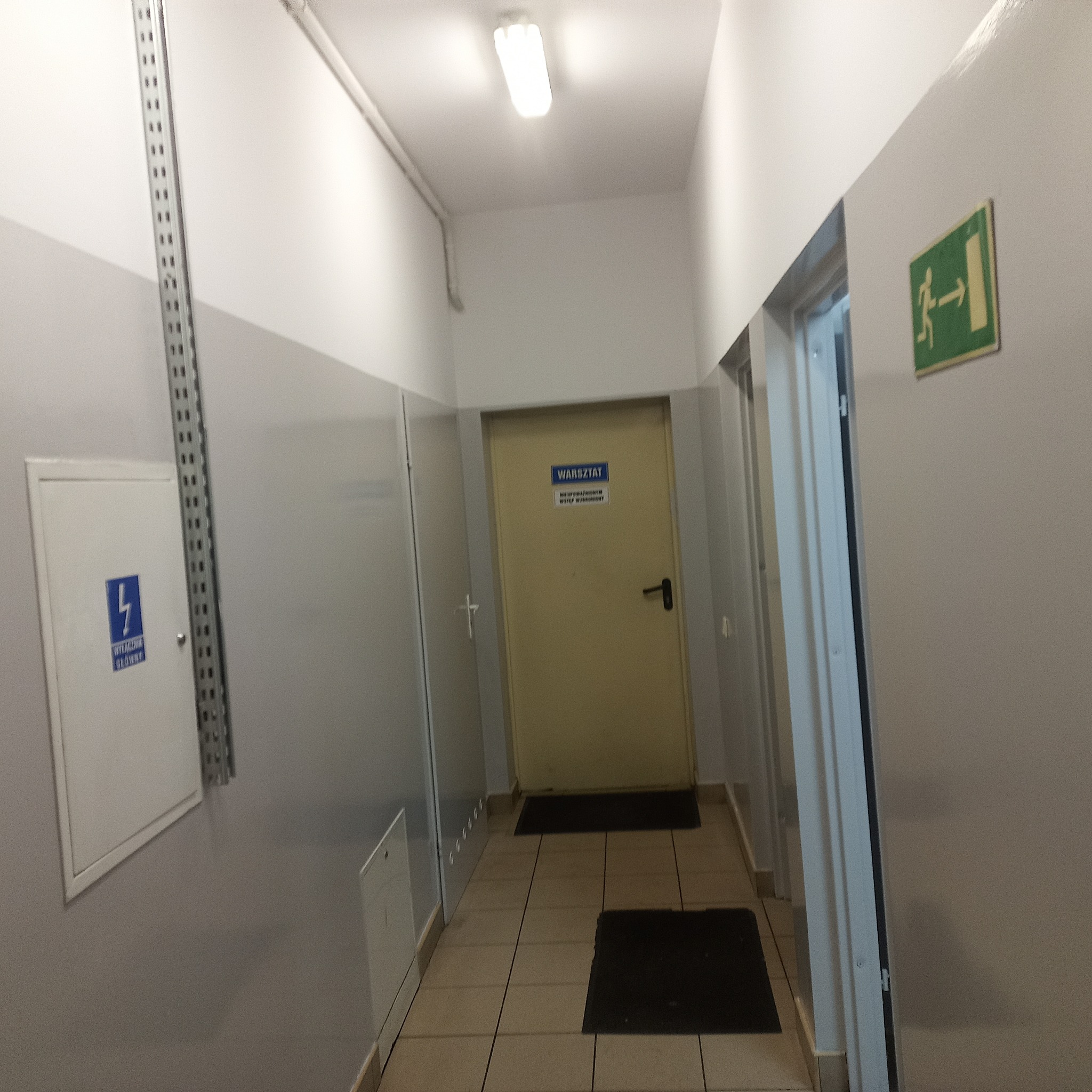 Korytarz w budynku z płytkami na podłodze, widoczne drzwi z napisem 'Warsztat', tabliczka z symbolem wyjścia ewakuacyjnego i skrzynka elektryczna.