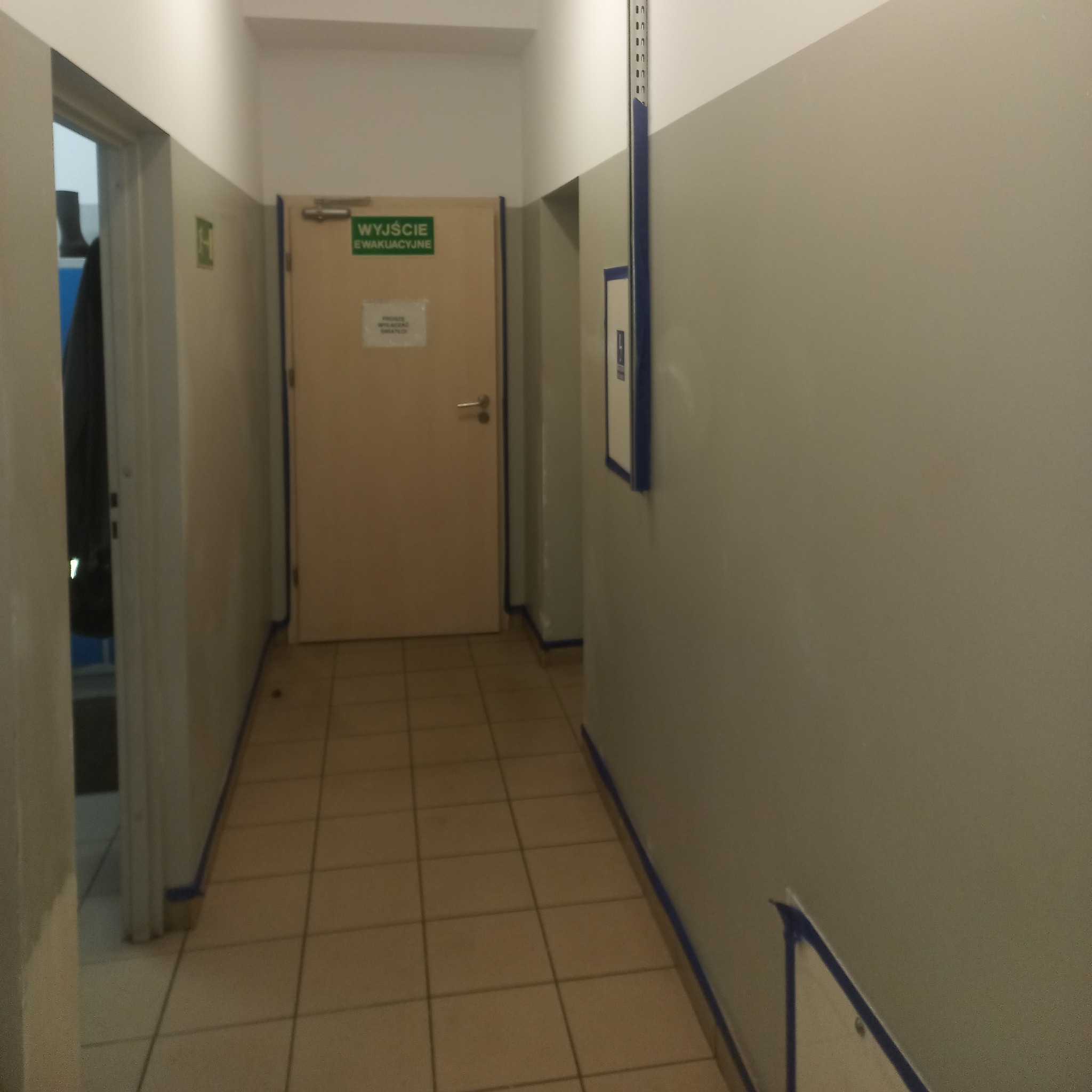 Korytarz biurowy w trakcie malowania, zabezpieczone taśmą malarską ściany i podłoga, widoczna tabliczka informacyjna 'Wyjście Ewakuacyjne'.