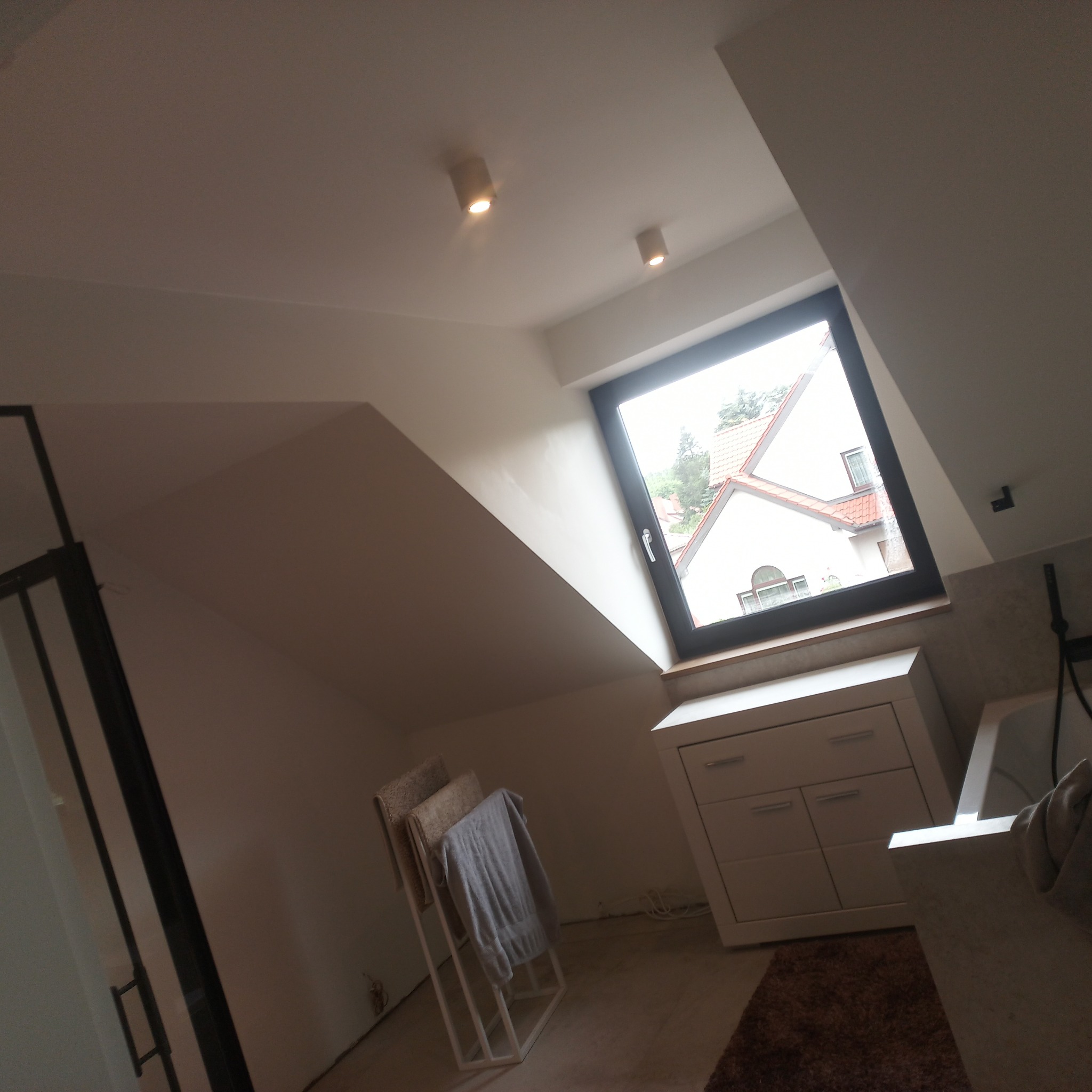 Wykończona łazienka na poddaszu z oknem dachowym, białą szafką pod umywalką, grzejnikiem drabinkowym na ręczniki, widoczne elementy instalacji wodnej.