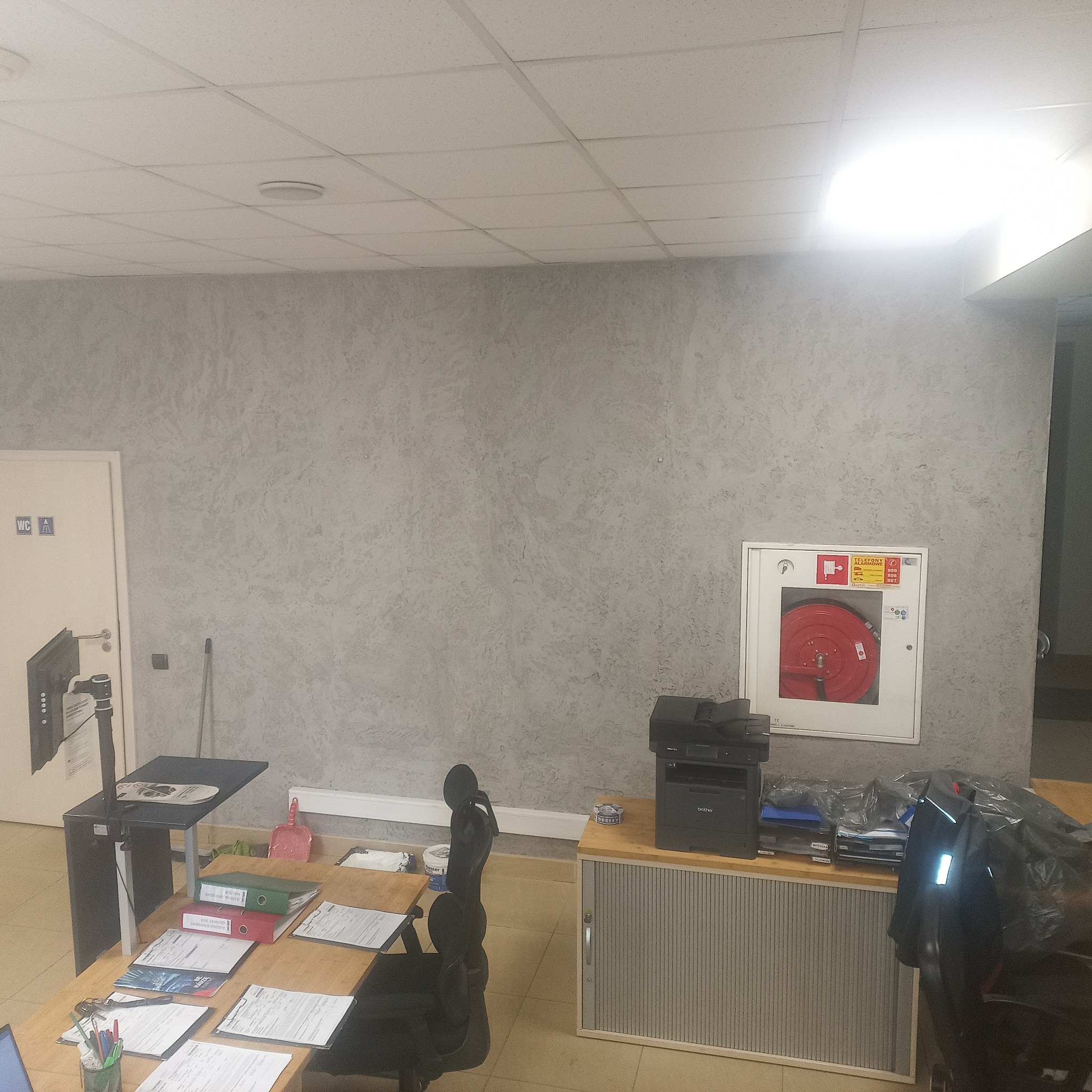 Biuro z szarą ścianą o fakturze baranka, biurkiem z dokumentami, monitorem, drukarką Brother, szafką z żaluzjami i hydrantem w białej obudowie.