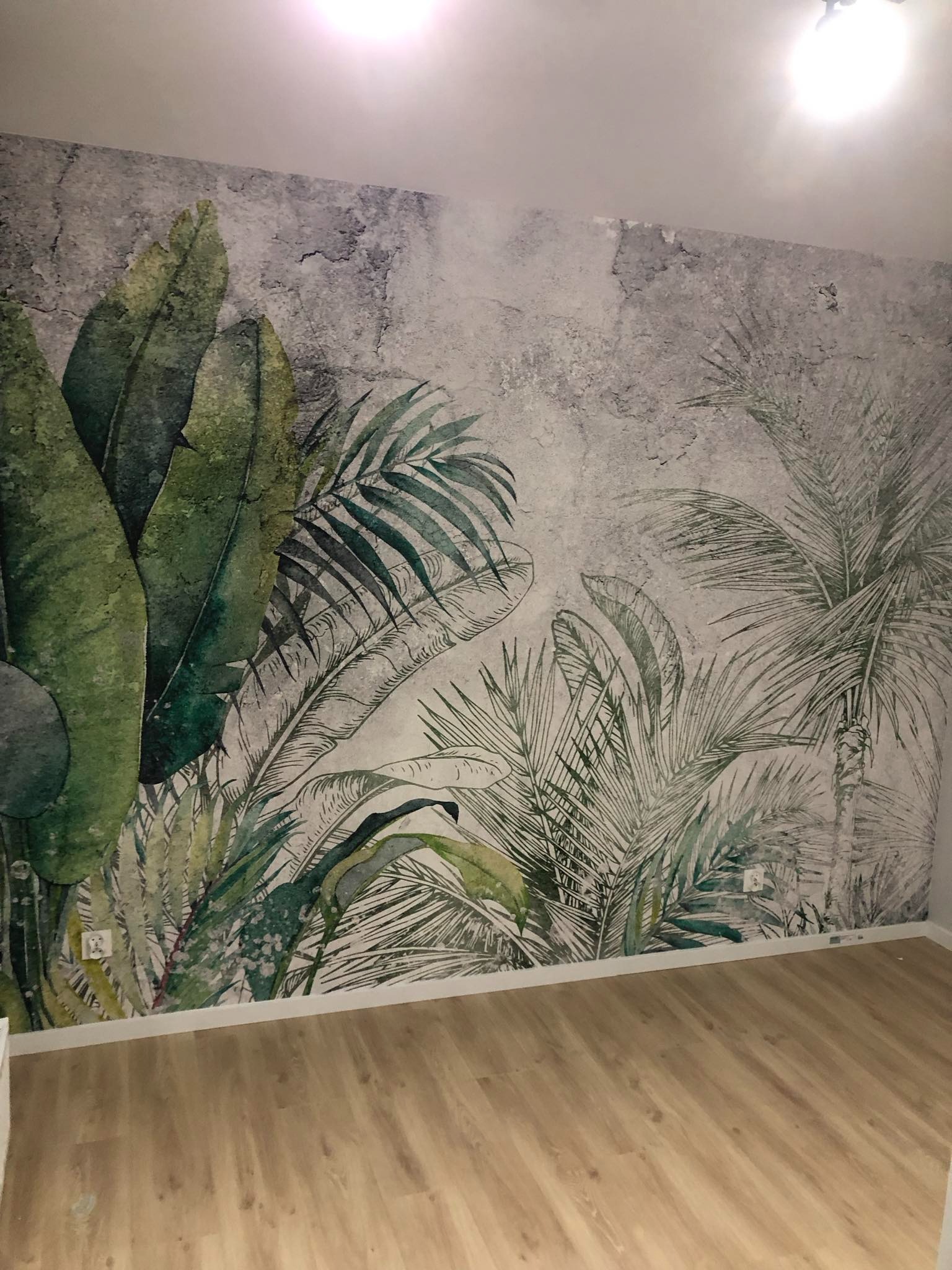 Pokój z fototapetą przedstawiającą motyw tropikalnych liści na teksturowanym, szarym tle, uzupełniony jasną podłogą z paneli i białymi listwami przypodłogowymi.