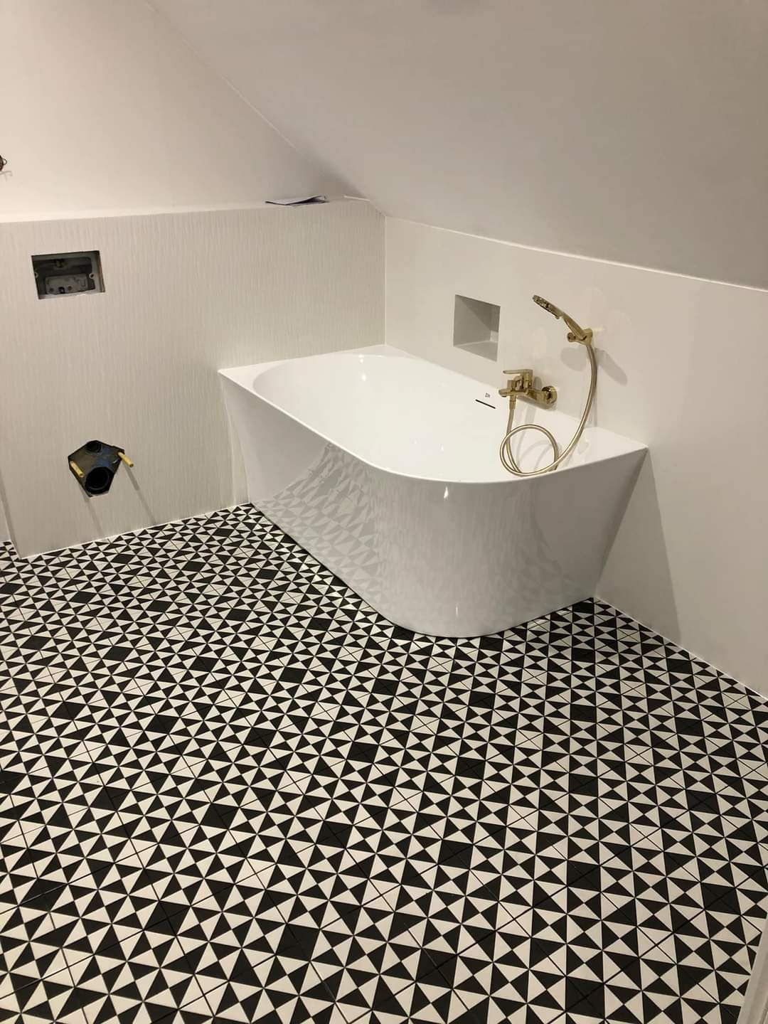 Nowoczesna łazienka na poddaszu z wolnostojącą wanną o nieregularnym kształcie, złotą armaturą i czarno-białą podłogą w geometryczny wzór. Widoczne niezabudowane otwory w ścianie.