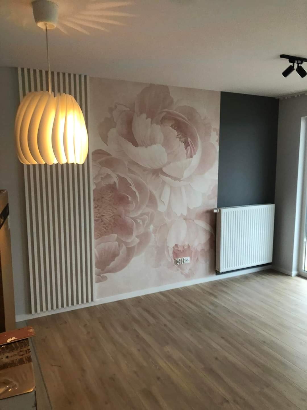 Pokój po odświeżeniu: ściana z fototapetą w pastelowe kwiaty, jasne panele podłogowe, biały grzejnik i designerska lampa wisząca.