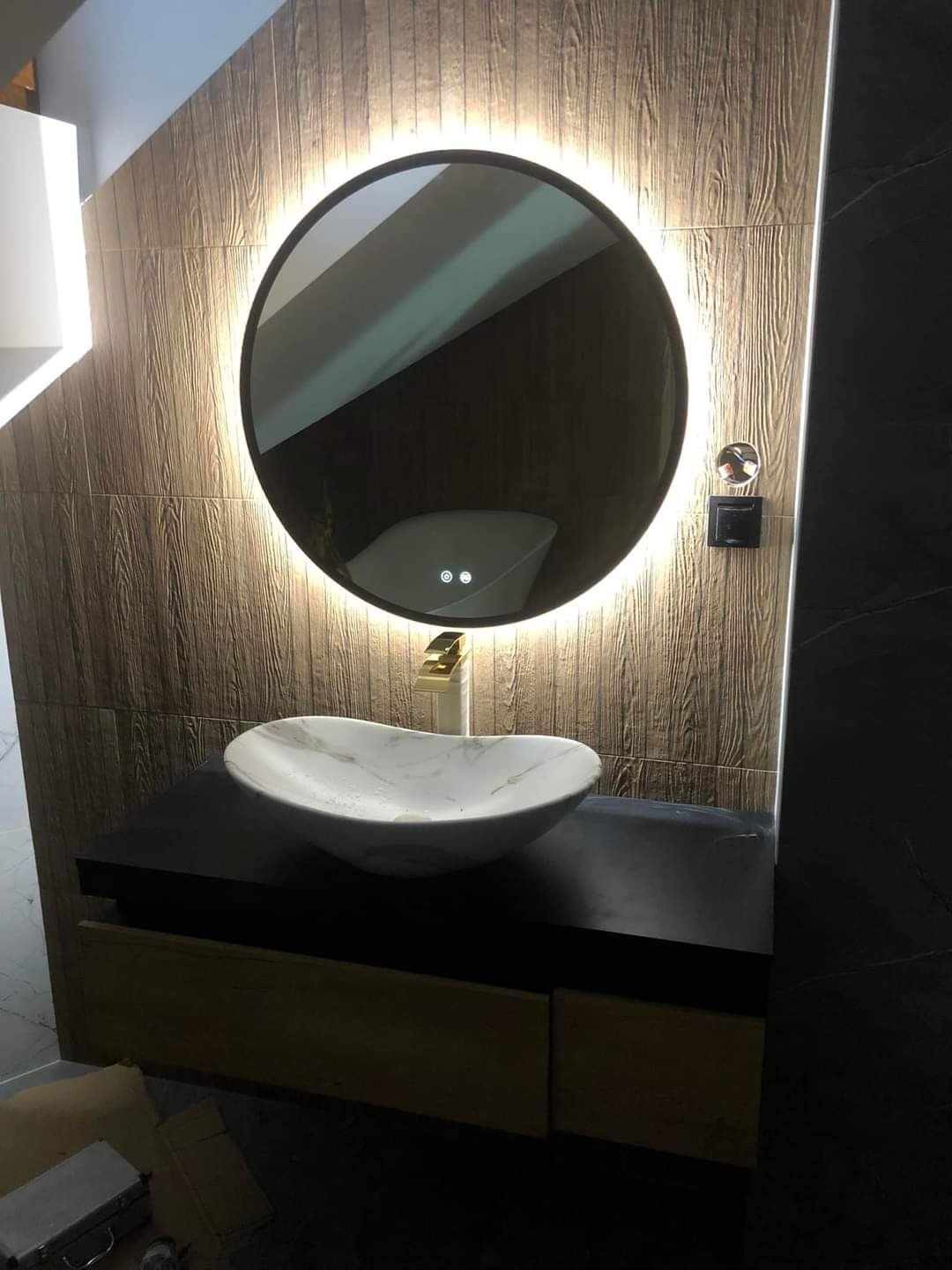 Nowoczesna łazienka z umywalką w kształcie łzy, okrągłym lustrem z podświetleniem LED oraz drewnopodobnymi płytkami na ścianie.