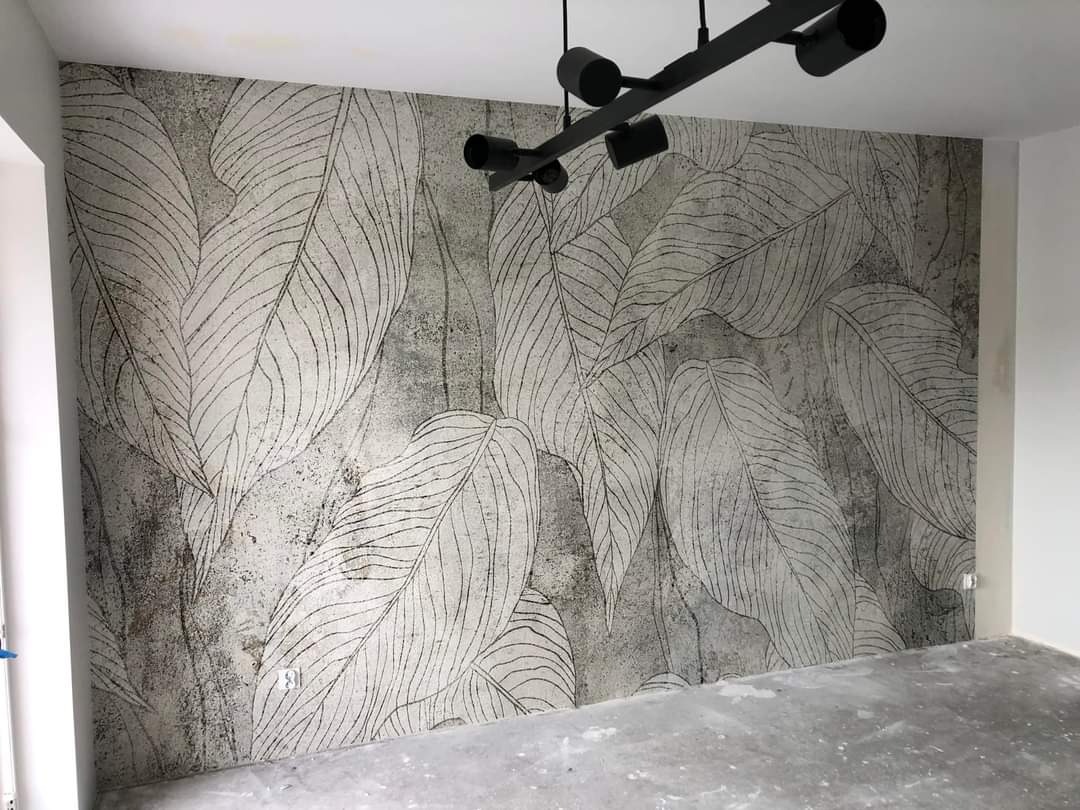 Ściana wykończona tapetą z motywem liści w odcieniach szarości, oświetlona nowoczesną czarną lampą sufitową, widoczny fragment surowej podłogi.