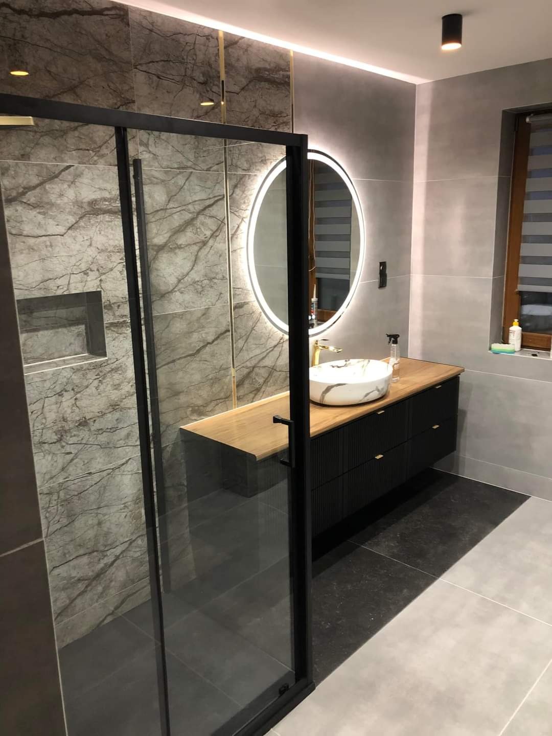 Nowoczesna łazienka z kabiną prysznicową typu walk-in, marmurowymi płytkami w strefie prysznica, okrągłym lustrem z podświetleniem LED, drewnianym blatem i czarną szafką pod umywalką.