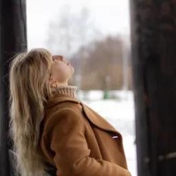 Kobieta w brązowym płaszczu opiera się o drewnianą barierkę, spoglądając w górę na zimowe niebo, widoczny fragment złotej kolczyka koła.