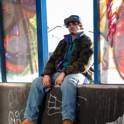 Jakub Tomkowicz - Młody mężczyzna w czapce siedzi na betonowym murku z graffiti, ubrany w jeansy, brązowe buty i kurtkę, z kolorowymi szybami za nim.