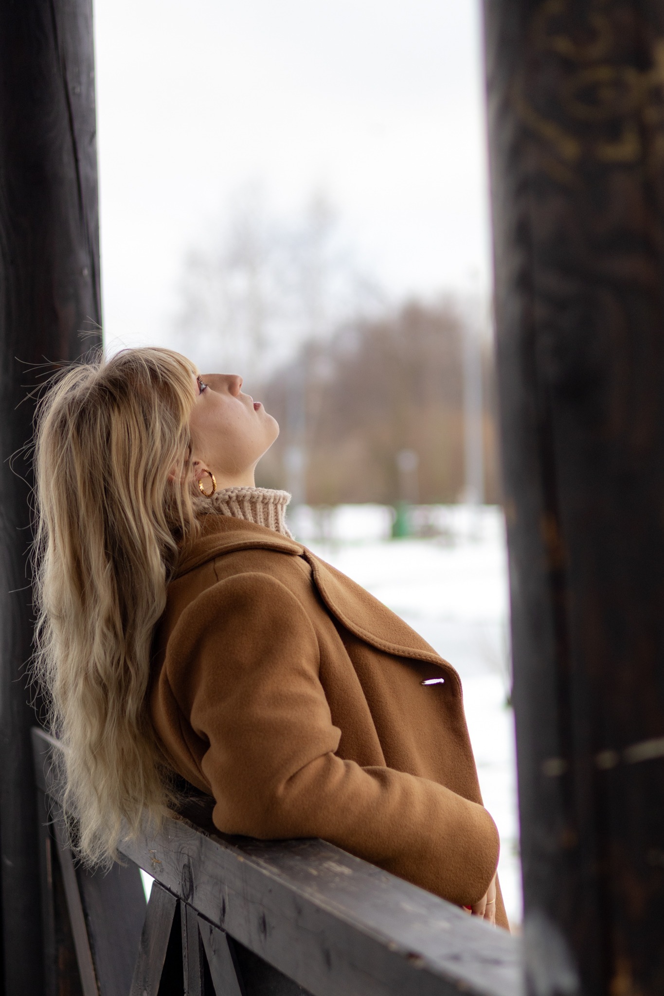 Kobieta w brązowym płaszczu opiera się o drewnianą barierkę, spoglądając w górę na zimowe niebo, widoczny fragment złotej kolczyka koła.