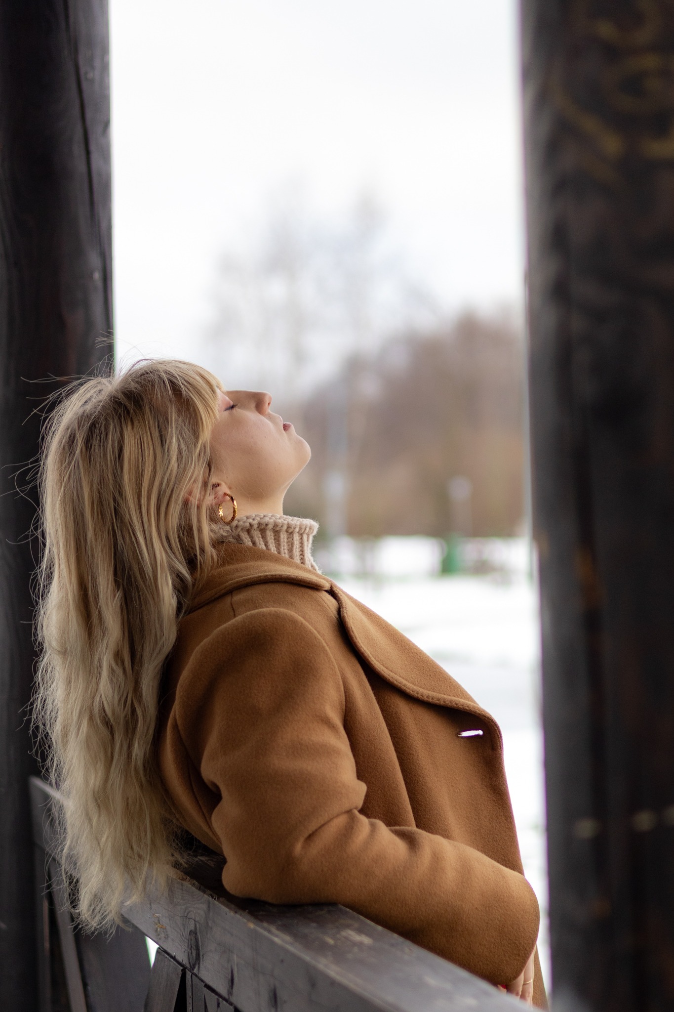 Kobieta w brązowym płaszczu opiera się o drewnianą barierkę, spoglądając w górę na zimowy krajobraz, widoczne złote kolczyki koła.