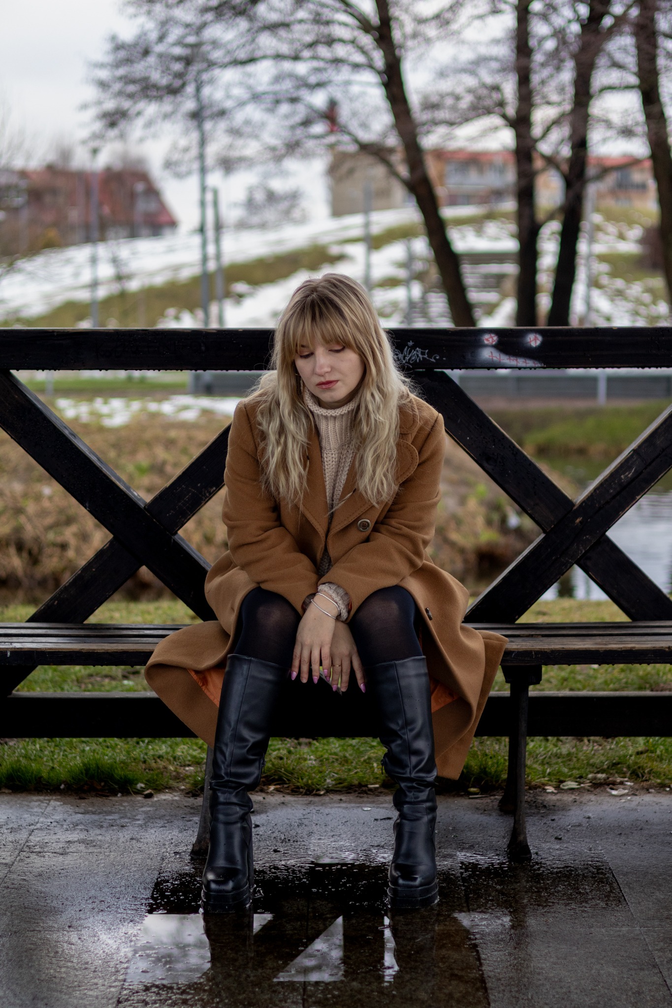 Zamyślona blondynka w brązowym płaszczu siedzi na czarnej ławce z graffiti, spoglądając w dół, w tle drzewa i budynki w zimowej scenerii.