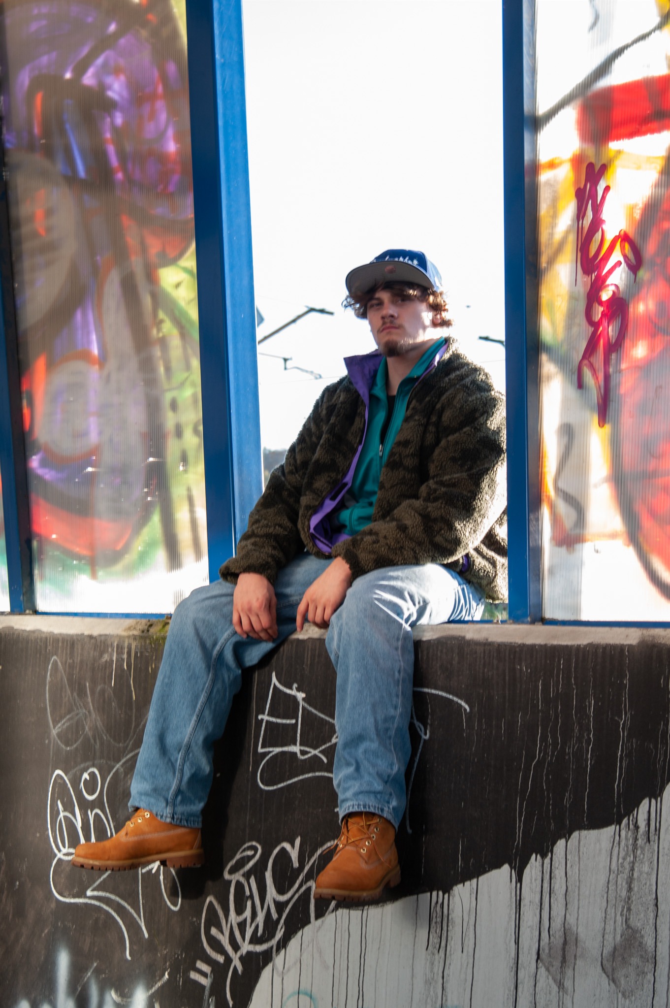 Młody mężczyzna w czapce siedzi na betonowym murku z graffiti, ubrany w jeansy, brązowe buty i kurtkę, z kolorowymi szybami za nim.