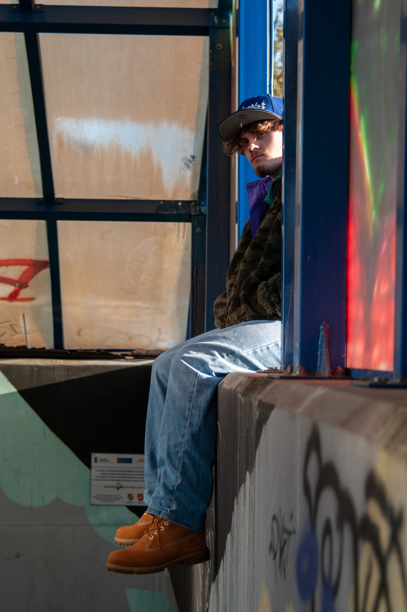 Młody mężczyzna w czapce i kurtce siedzi na betonowej ścianie przystanku autobusowego z graffiti, nosząc dżinsy i brązowe buty.
