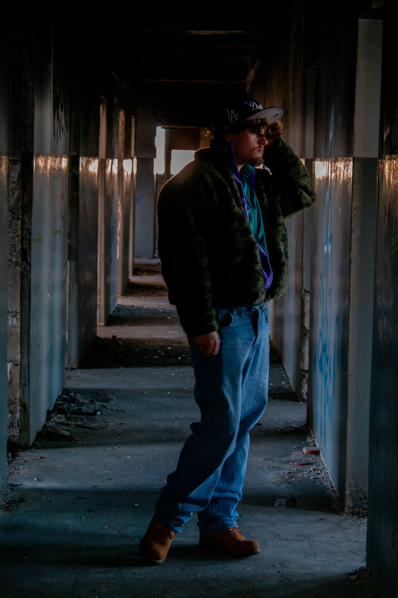 Mężczyzna w czapce z daszkiem i kurtce w stylu vintage pozuje w opuszczonym korytarzu z graffiti.
