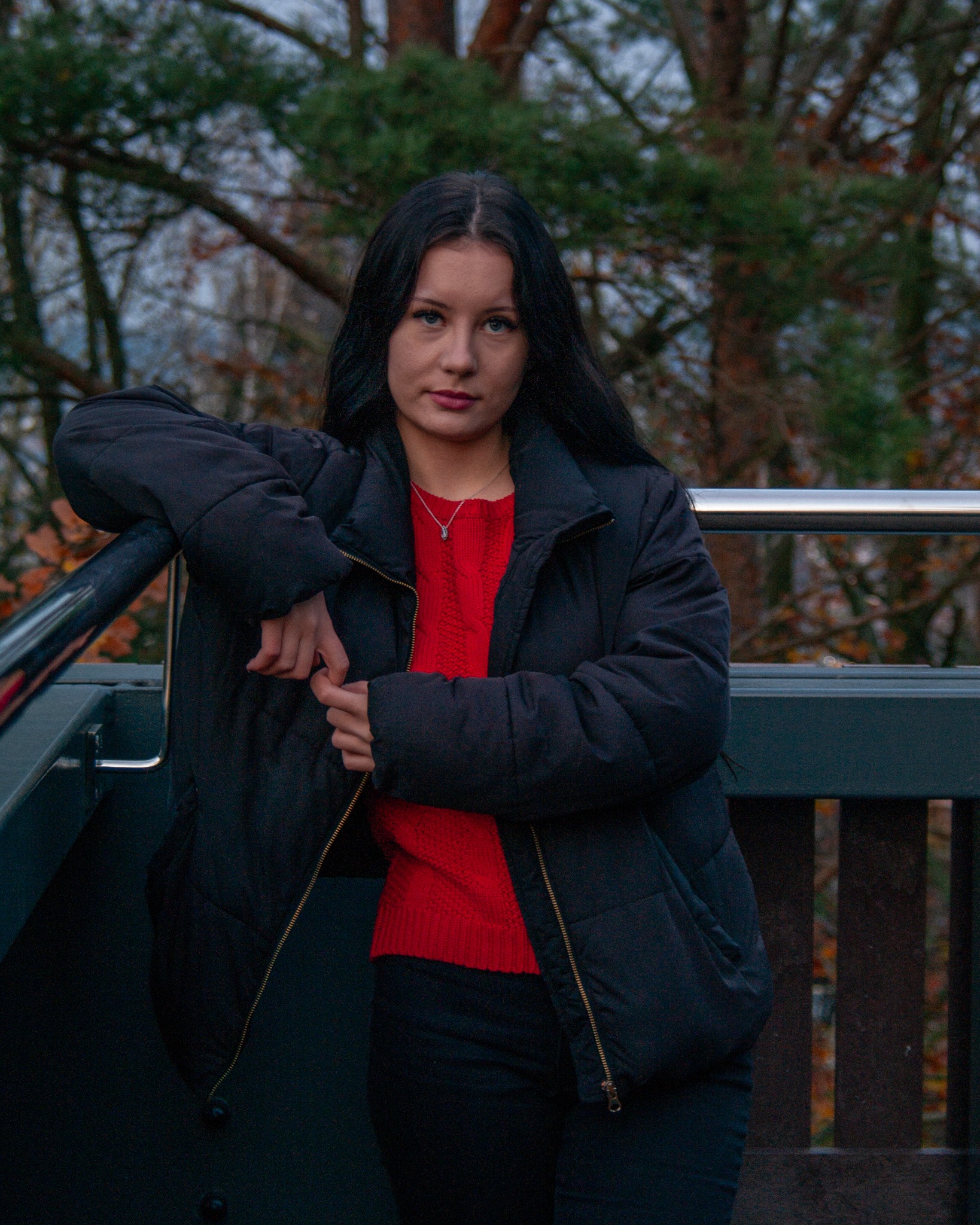Portret młodej kobiety z długimi, czarnymi włosami, ubranej w czerwoną bluzkę i czarną kurtkę, opierającej się o metalową barierkę na tle drzew.