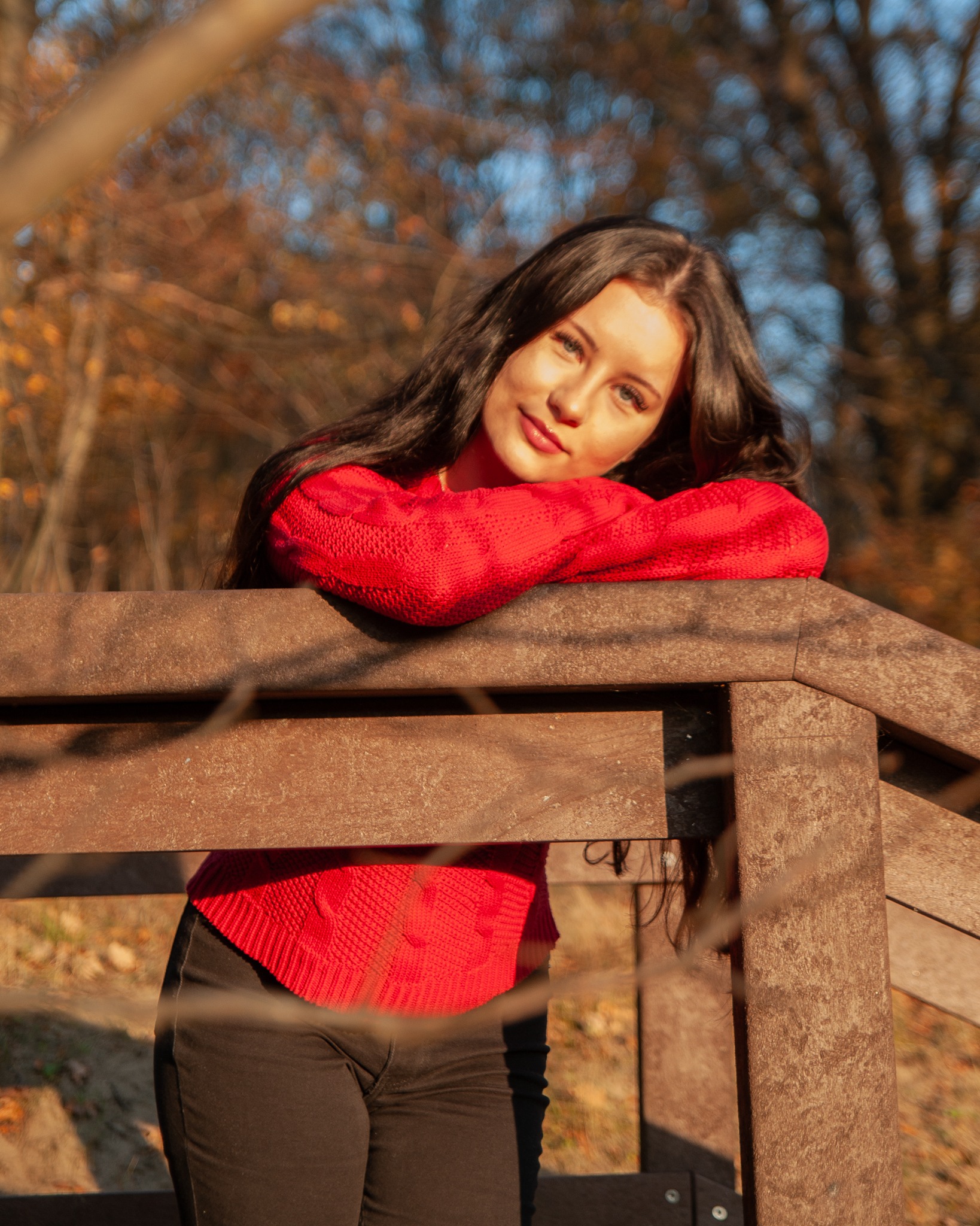 Portret młodej kobiety w czerwonym swetrze opierającej się o drewnianą barierkę na tle jesiennego lasu z rozproszonym światłem.