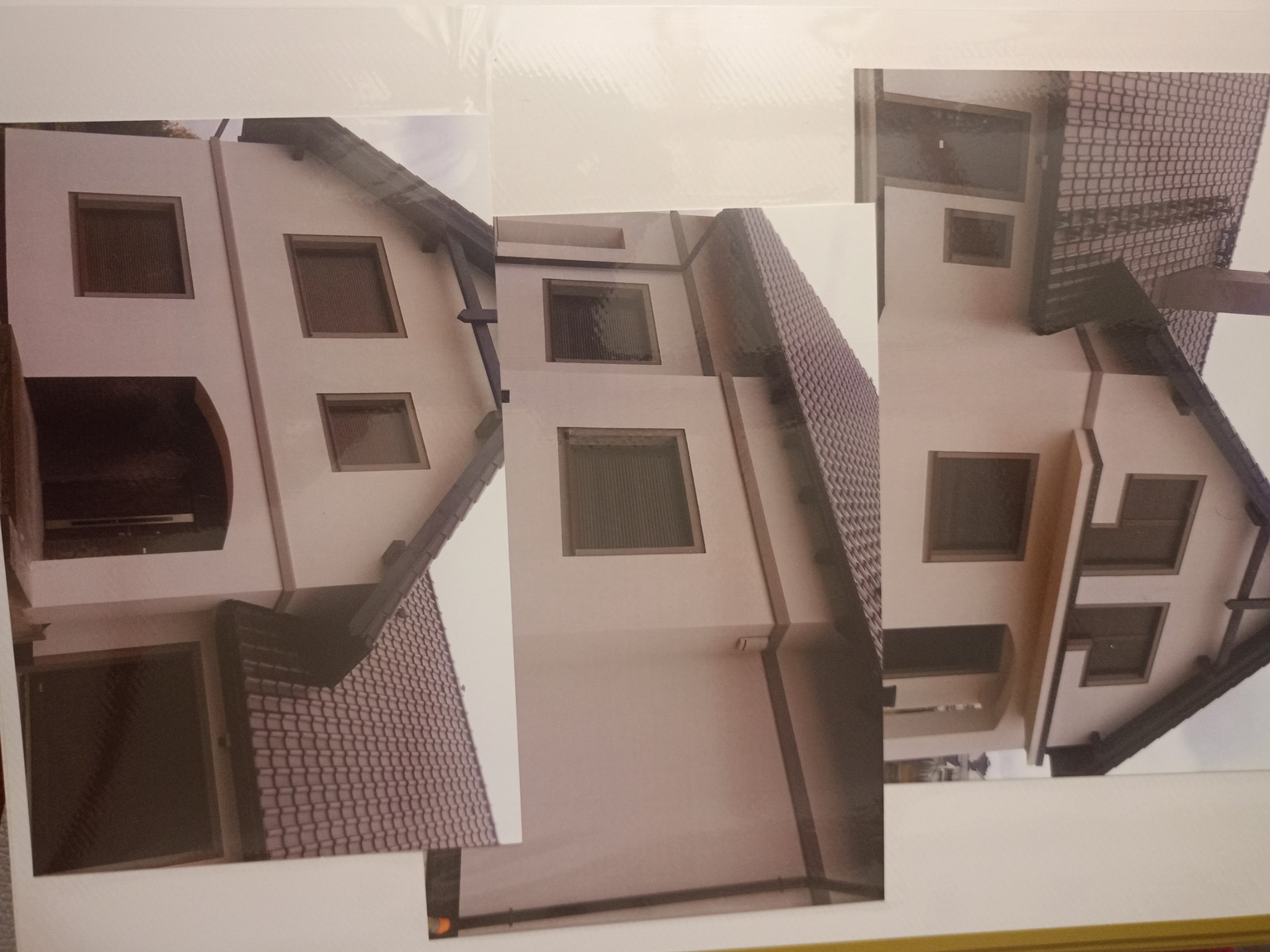 Zdjęcie fragmentów elewacji domu z oknami, wydrukowane na papierze i ułożone obok siebie, tworząc kolaż. Widoczna jasna elewacja, ciemna dachówka i białe tło papieru.