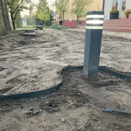 Przygotowany teren pod ogród z wytyczonymi rabatami obrzeżami z tworzywa sztucznego, widoczna lampa ogrodowa i przygotowane podłoże. W tle budynek mieszkalny i drzewa.