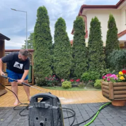 Mężczyzna w czarnej koszulce z logo Adidas i niebieskich spodenkach czyści kostkę brukową myjką ciśnieniową na podwórku z tujami i kwiatami w drewnianej donicy.
