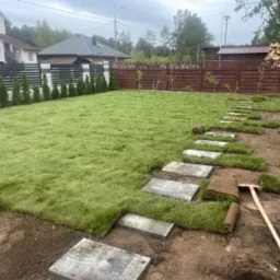 Układanie trawy z rolki i ścieżki z płyt betonowych w ogrodzie, widoczne narzędzie ogrodnicze i częściowo rozłożone rolki trawy.