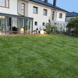 P.H.U Konrad Pacholczyk Metrycki - Świeżo założony trawnik z rolki przed nowoczesnym domem z przeszkloną werandą. Dekoracyjna kula świetlna i osoba z koszem kwiatów dodają uroku.